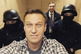 Alexei Navalny smiles for the camera while two men in black police uniforms stand behind him.