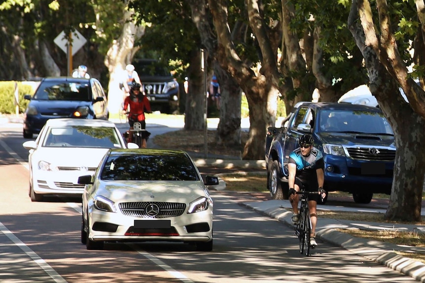 A white car overtakes a cyclist on a suburban road, with other vehicles travelling behind.