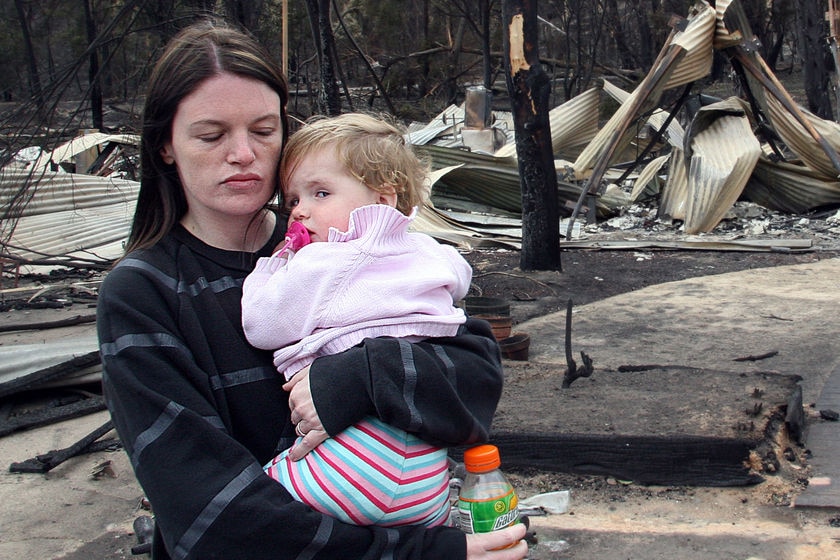 A woman, with eyes closed, holds her baby niece in front of her bushfire-destroyed home