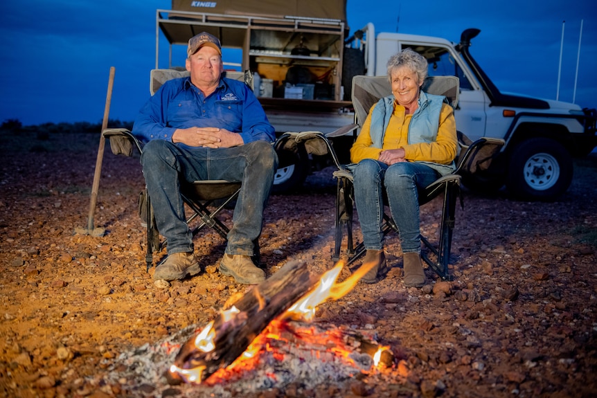 Man and women sitting in camp chairs behind a campfire at night in the bush with a Ute parked behind them. 