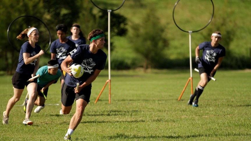 A man dressed in a team sporting uniform runs with a ball, hoops on poles and female and male players in background.