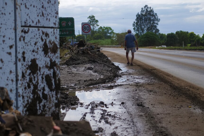 A man walks along a muddy road which has a flood-damaged and mud-speckled fridge.