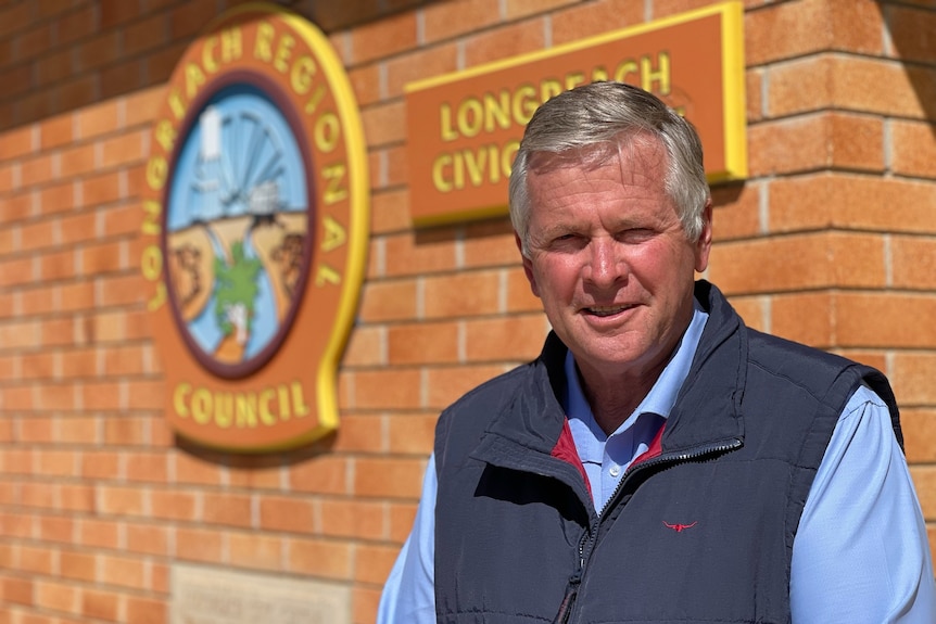 An older man stands in front of the Longreach Regional Council sign, wearing a dark button-up and looking apprehensive.