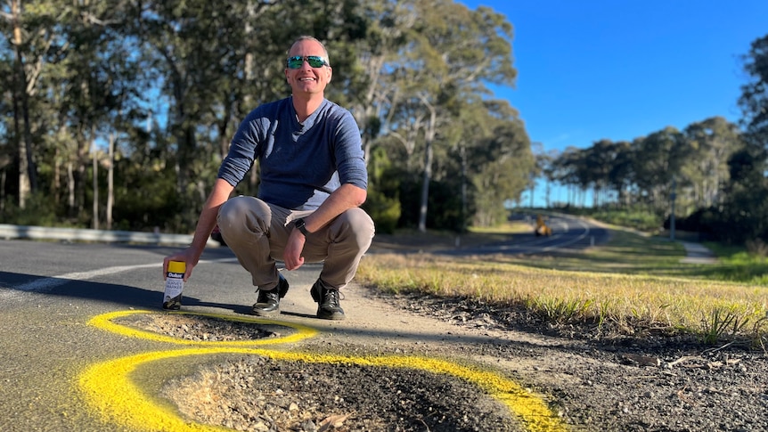 A man squats in front of two big potholes holding a spray can.