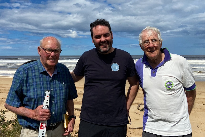 Three men shoulder to shoulder with a beach in the background.