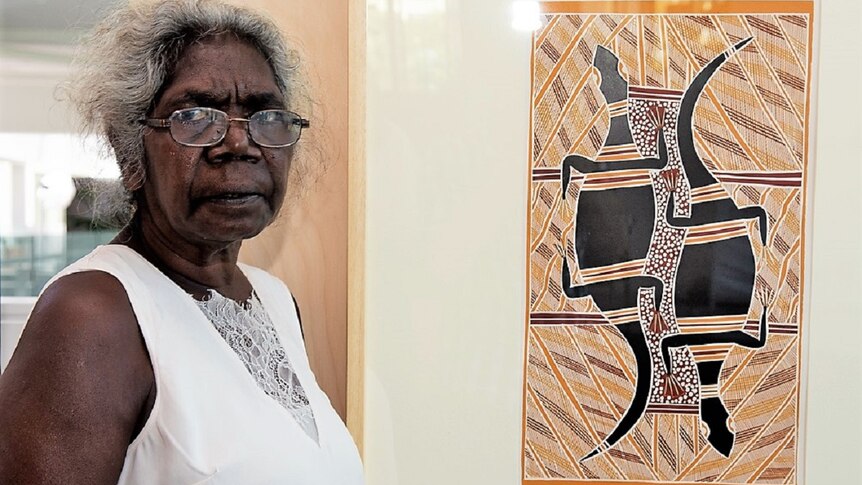 An Aboriginal woman stands in front of a painting.