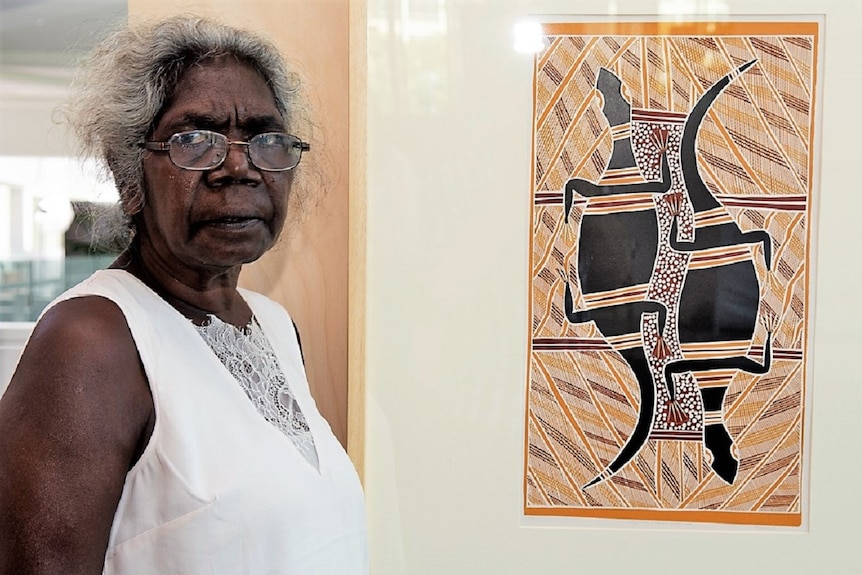 An Aboriginal woman stands in front of a painting.