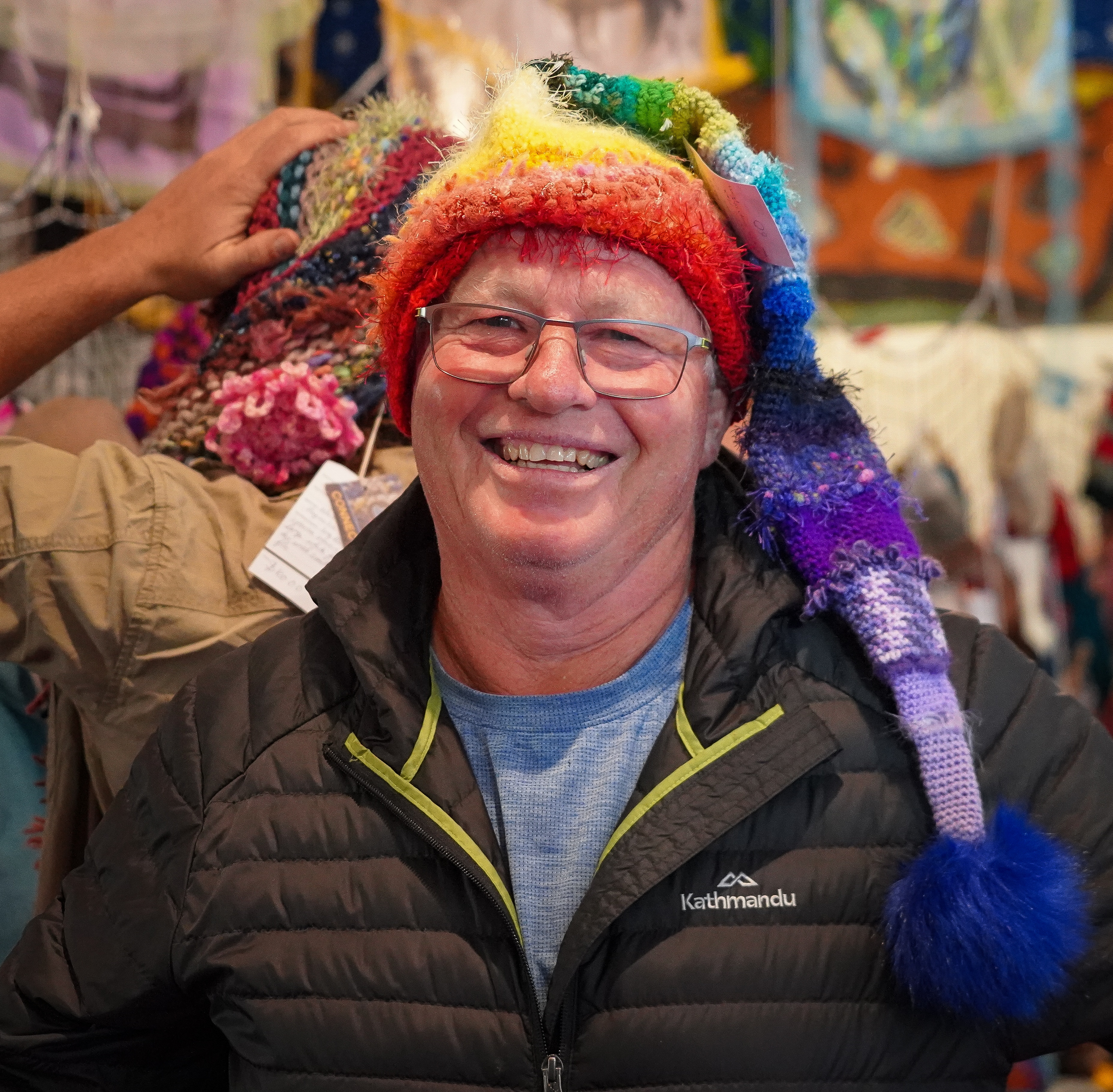 A man in reading glasses smiles at the camera, he is wearing a rainbow-coloured knitted beanie.