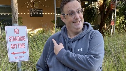 man with spectacles in blue hoodie sitting in wheelchair pointing at 'no standing' sign