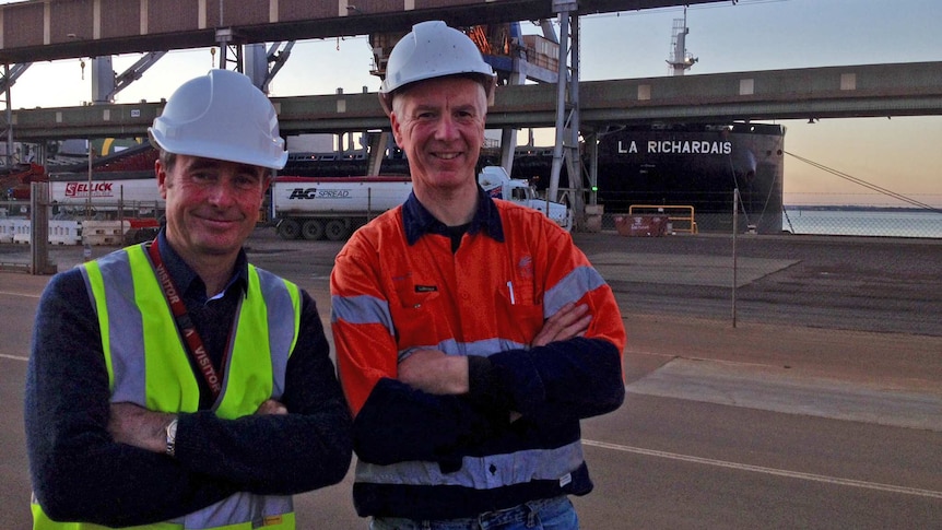 Two men, stand crossed armed, in high visibility clothing with a ship behind them, loading wood chips.