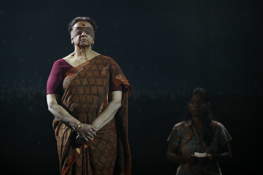 A Sri Lankan woman in her mid 60s wearing a blindfold and brown and red sari, stands arms clasped