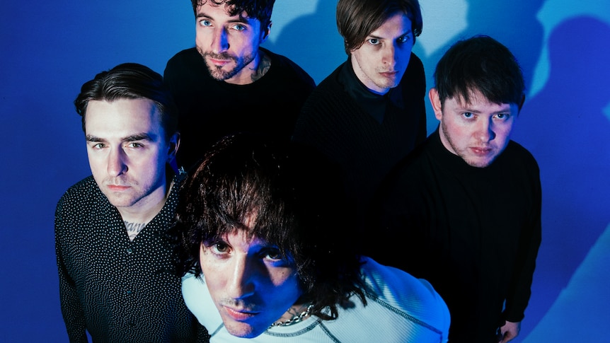 The five members of Bring Me The Horizon stand together against a blue background and look up at the high angle camera