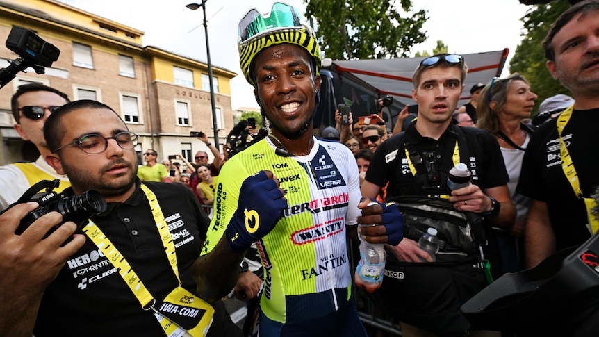 A Black African cyclist stands smiling with fist pumped at the end of a stage in the Tour de France.