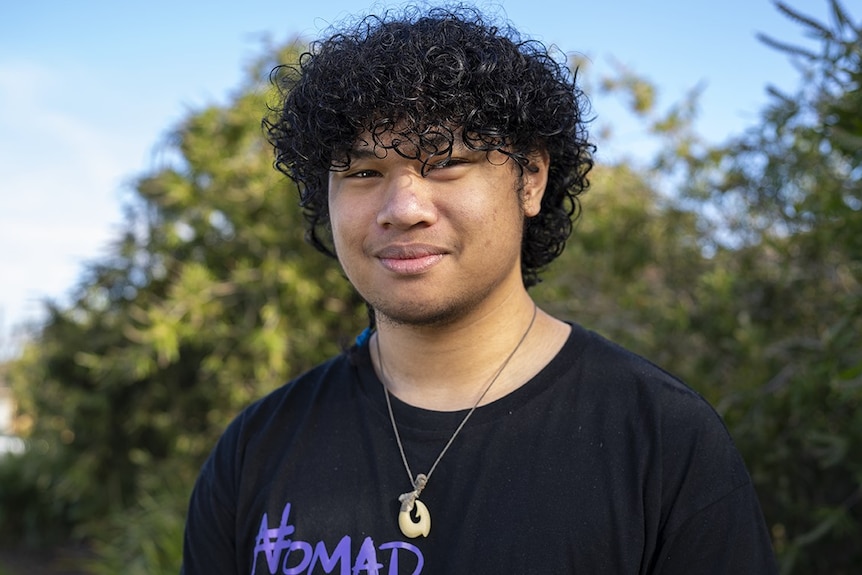 A man with curly black hair smiles at the camera. he stands in front of a suburban home.