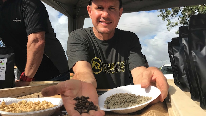 Man sits at market stall holding coffee beans in the palm of his outstretched hand.