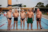 Five men in swim-wear stand at the end of an outdoor swimming pool looking in different directions and smiling.