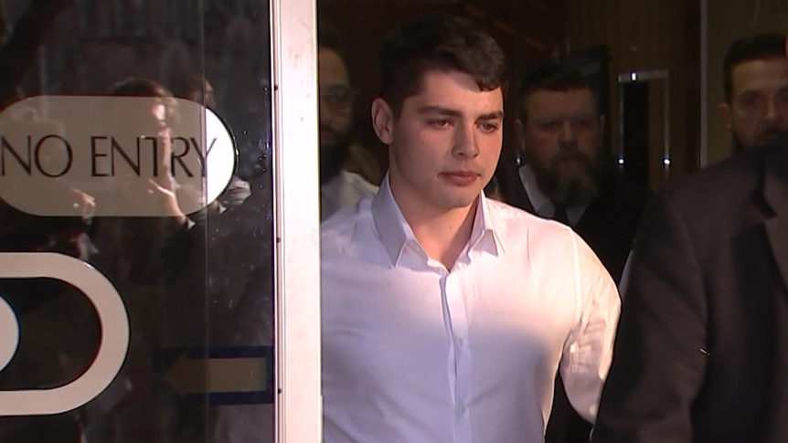 A young man in a white shirt walks through sliding glass doors surrounded by people