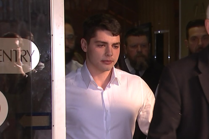 A young man in a white shirt walks through sliding glass doors surrounded by people