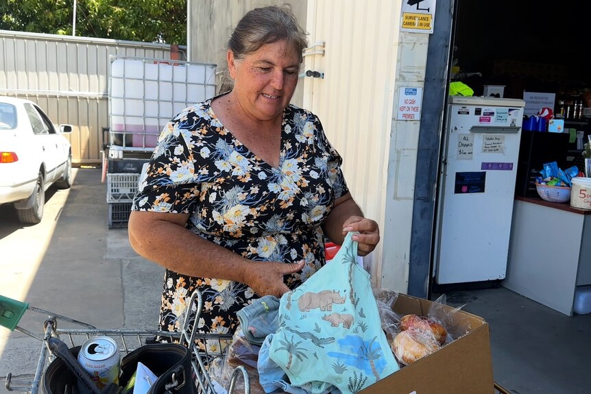 A woman with grey hair wearing a dress in front of a trolley with food and baby clothes