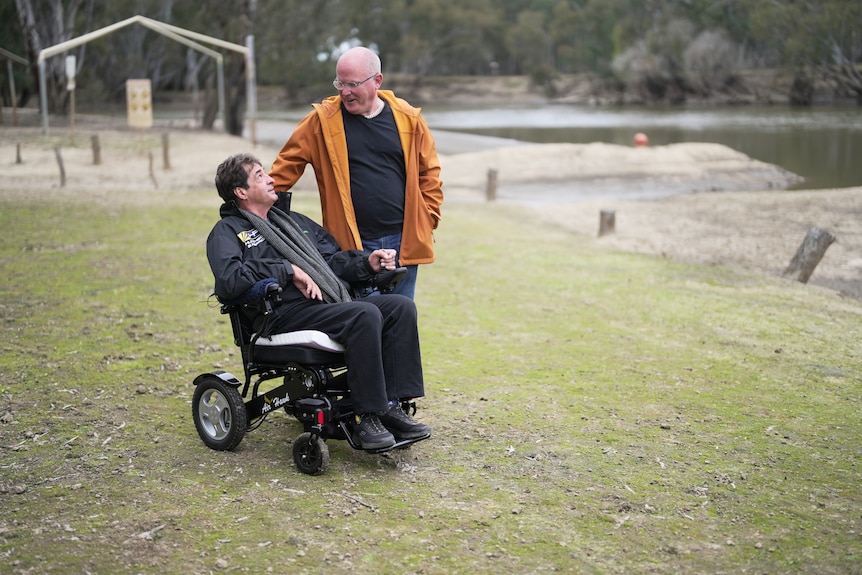 A man in a wheelchair looks up at another man who is standing next to him outside 