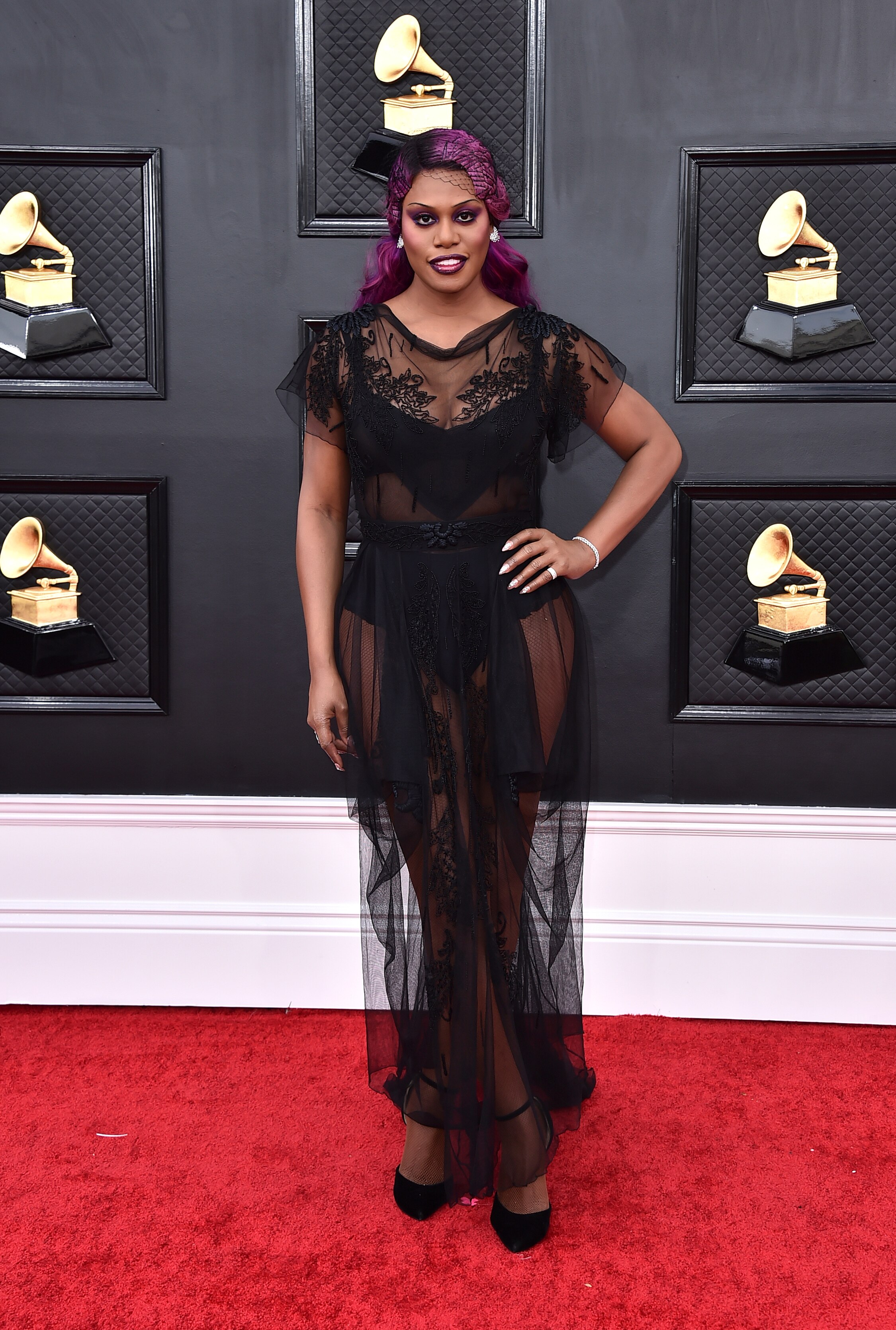 laverne cox poses on the grammys red carpet wearing a sheer black dress with lace accents and a black mesh headpiece