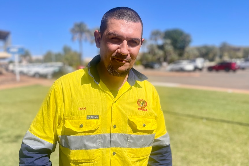A man stares sternly at the camera with his hands on his hips in a high vis shirt. Background is blurred. 