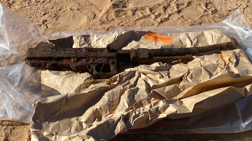 The rusty metal body of gun sitting on paper and tarp in sand.