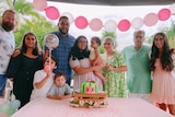 A family photo of parents and children standing around a cake with pink balloons above