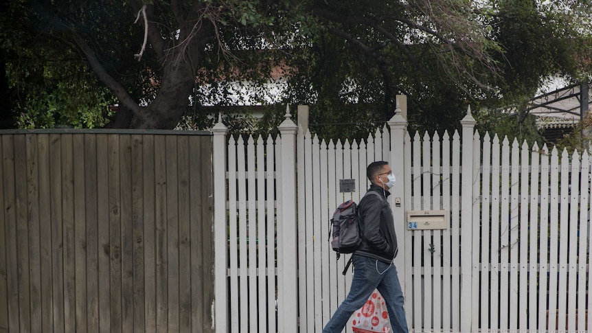 A man wearing a face mask walks on a residential street holding a shopping bag.