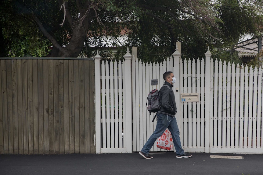 A man wearing a face mask walks on a residential street holding a shopping bag.