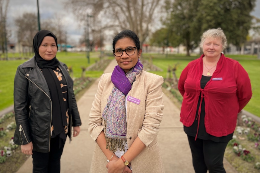 Three women stand on a pathway in a park.