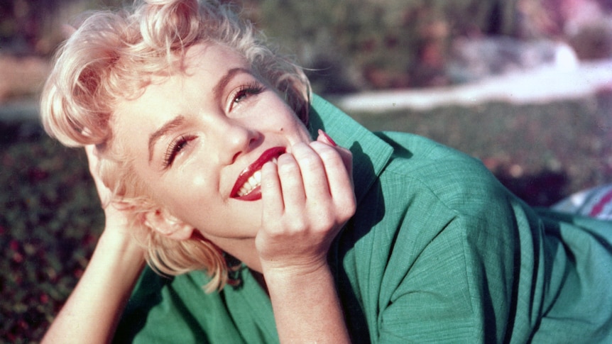 Marilyn Monroe lays on her stomach on a lawn, smiling into the distance as she rests her chin on her right hand