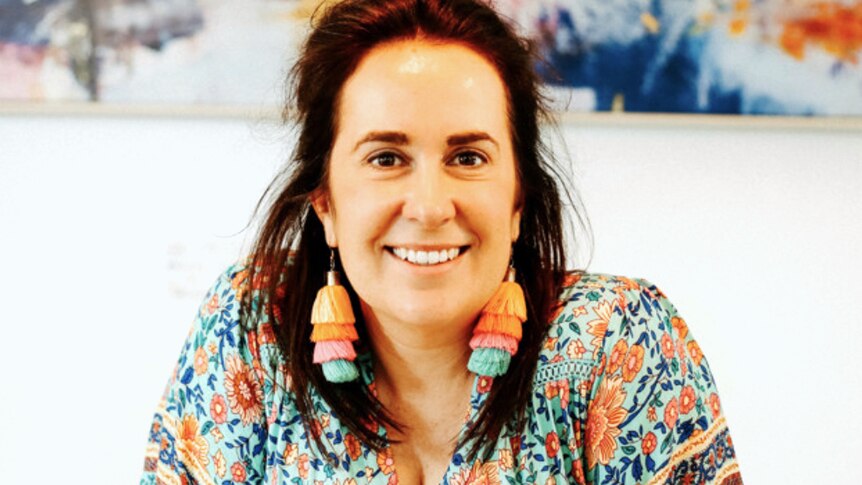Lucy Moss sits a table with colourful earrings and shirt, with painting in the background.
