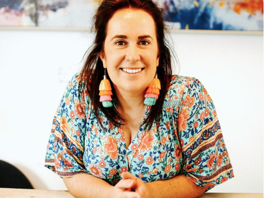 Lucy Moss sits a table with colourful earrings and shirt, with painting in the background.