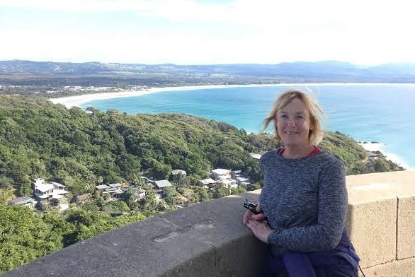 A blond woman standing at a brick wall overlooking green hills and the ocean.