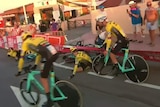 Cyclists in yellow jerseys ride past teammates who are lying on the ground next to a crash barrier