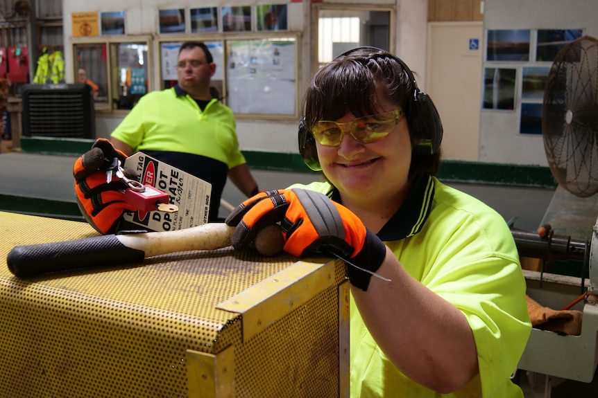 A woman in high-visibility clothing leans rests her hands on a yellow machine and smiles facing the camera