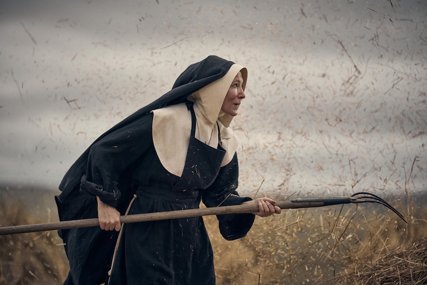 Actress Cate Blanchett, dressed as a nun, standing in a windy field holding a pitchfork.