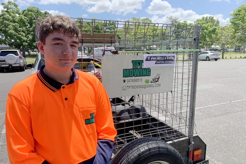 Teenager in high vis orange T-shirt standing in front of a trailer with a mower in it