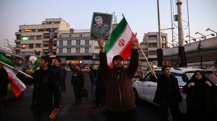 A man holds Qassem Soleimani's picture up as people on both sides hold Iranian flags. They are wearing dark colours, some shout.