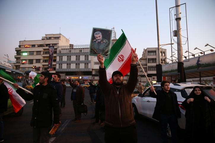 A man holds Qassem Soleimani's picture up as people on both sides hold Iranian flags. They are wearing dark colours, some shout.