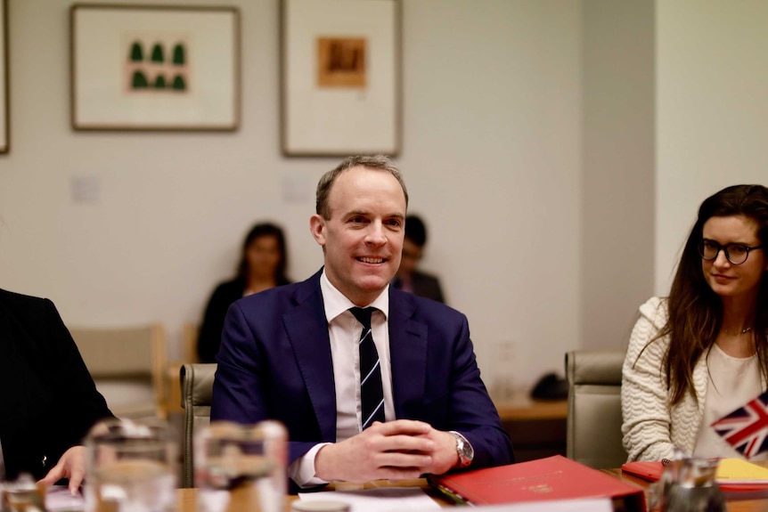 Dominic Raab sits at a table with his hands together. He is surrounded by staff.