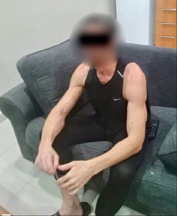 A lean man wearing exercise clothes sits on a couch, with his faced blurred to prevent identification.