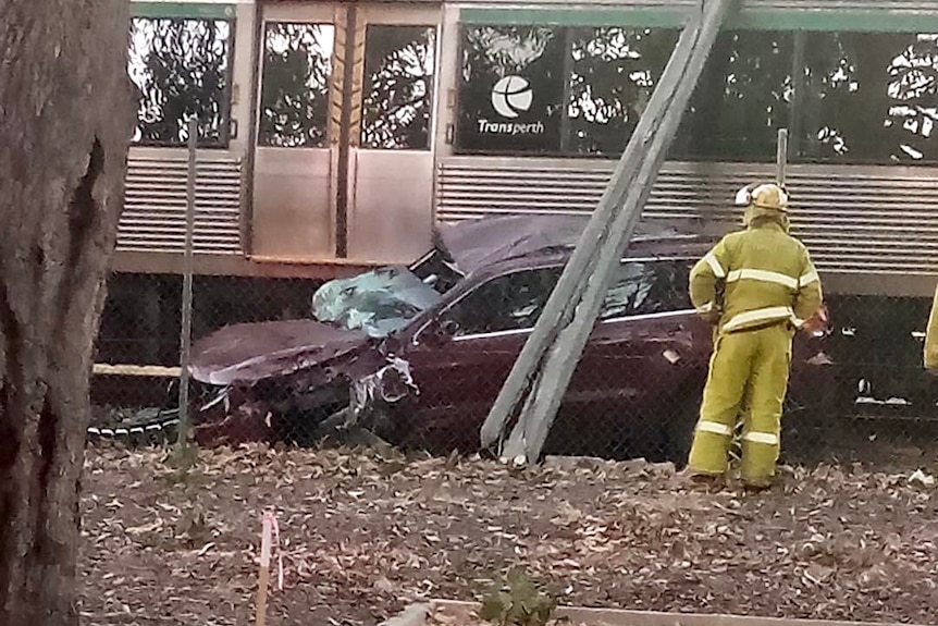 A side-on shot of the wreckage of a dark red car crushed net to a train with a firefighter standing nearby in yellow clothing.