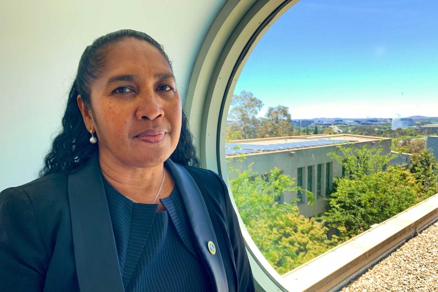 Vonda Malone wears a navy suit and stands in front of a circular window with blue sky, green trees and solar panels outside.