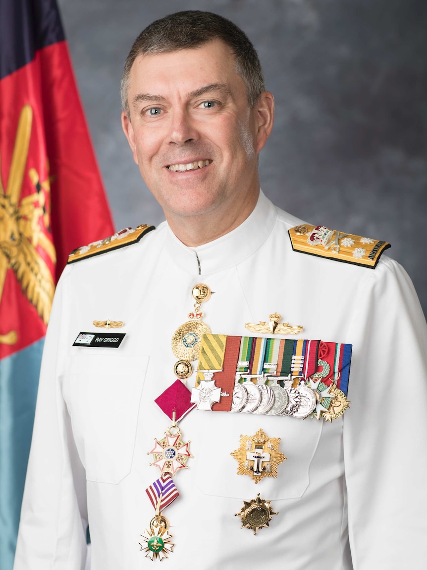 Ray Griggs, wearing his Air Force uniform, sits in front of a grey background and Australian flag.