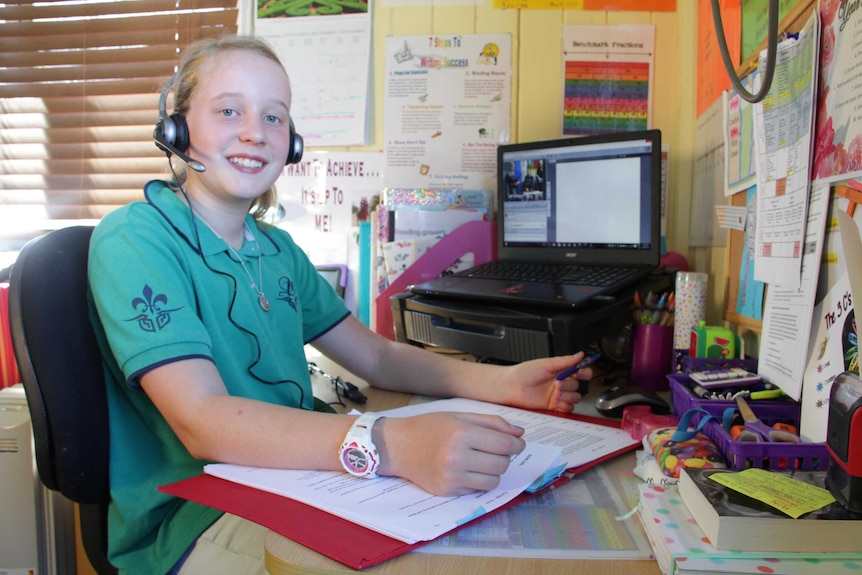 A girl wearing headphones and microphone sits at a school desk.