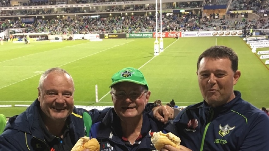 Three men in Canberra Raiders merchandise stand in the stands of a rugby stadium