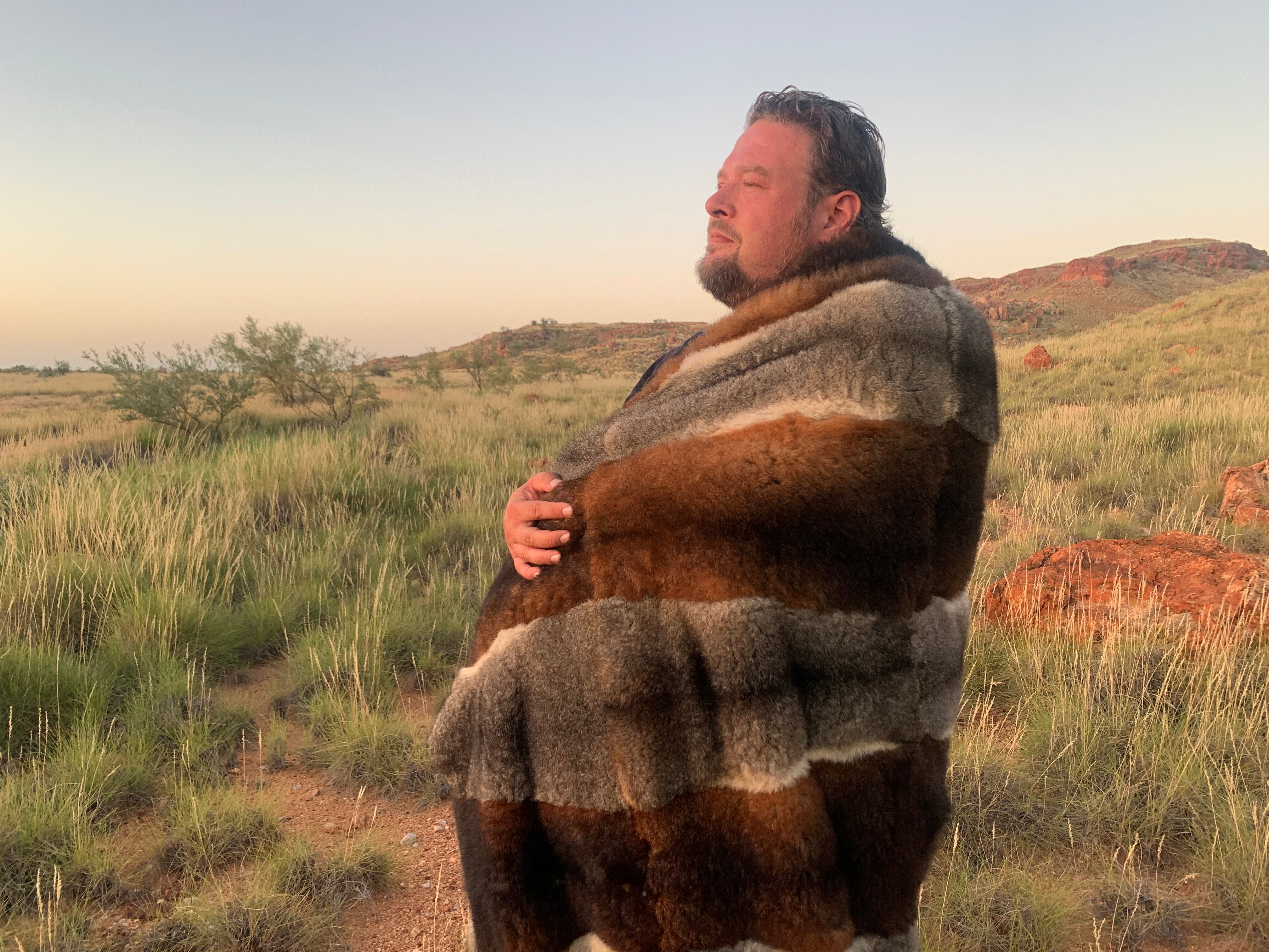 A tall Aboriginal man, wrapped in an opossum skin coat, stands in the outback