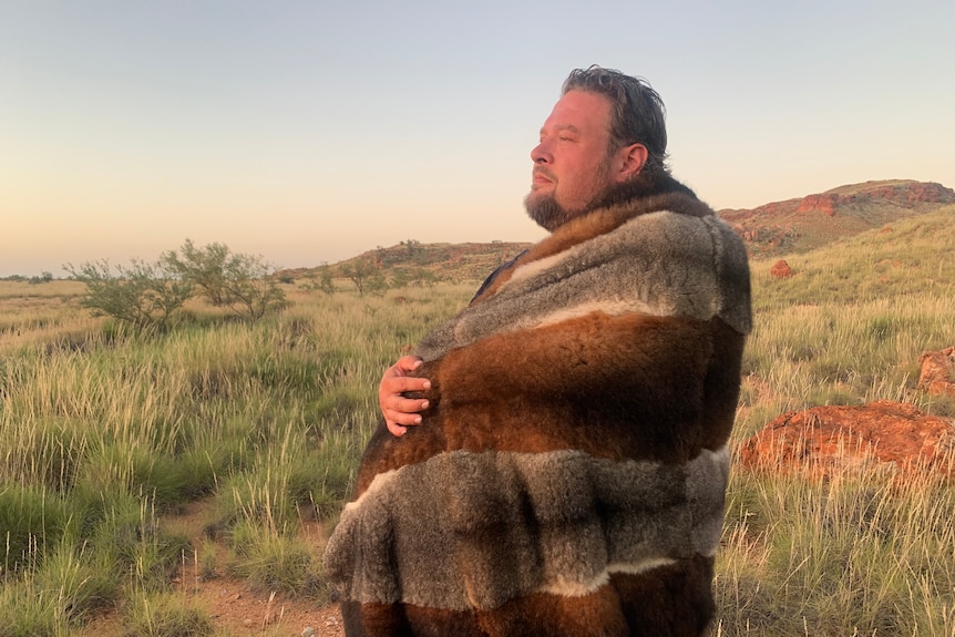 A tall Aboriginal man, wrapped in an opossum skin coat, stands in the outback